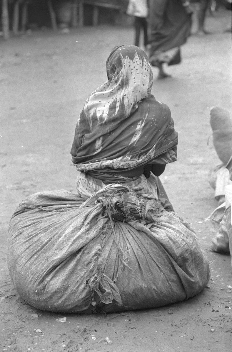 A woman seen from behind, sitting on a large tied-up sack on a dusty street.