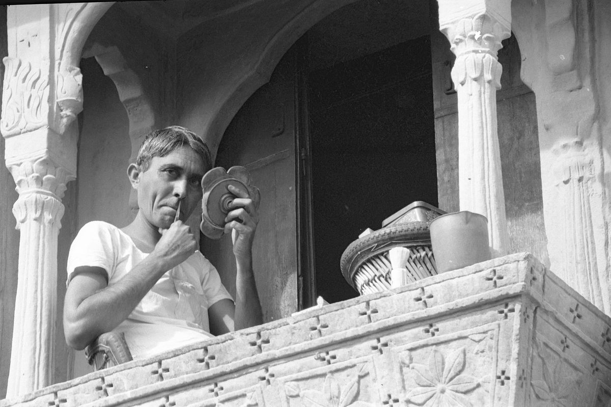 A man in a vest shaving at a mirror on a carved stone balcony.