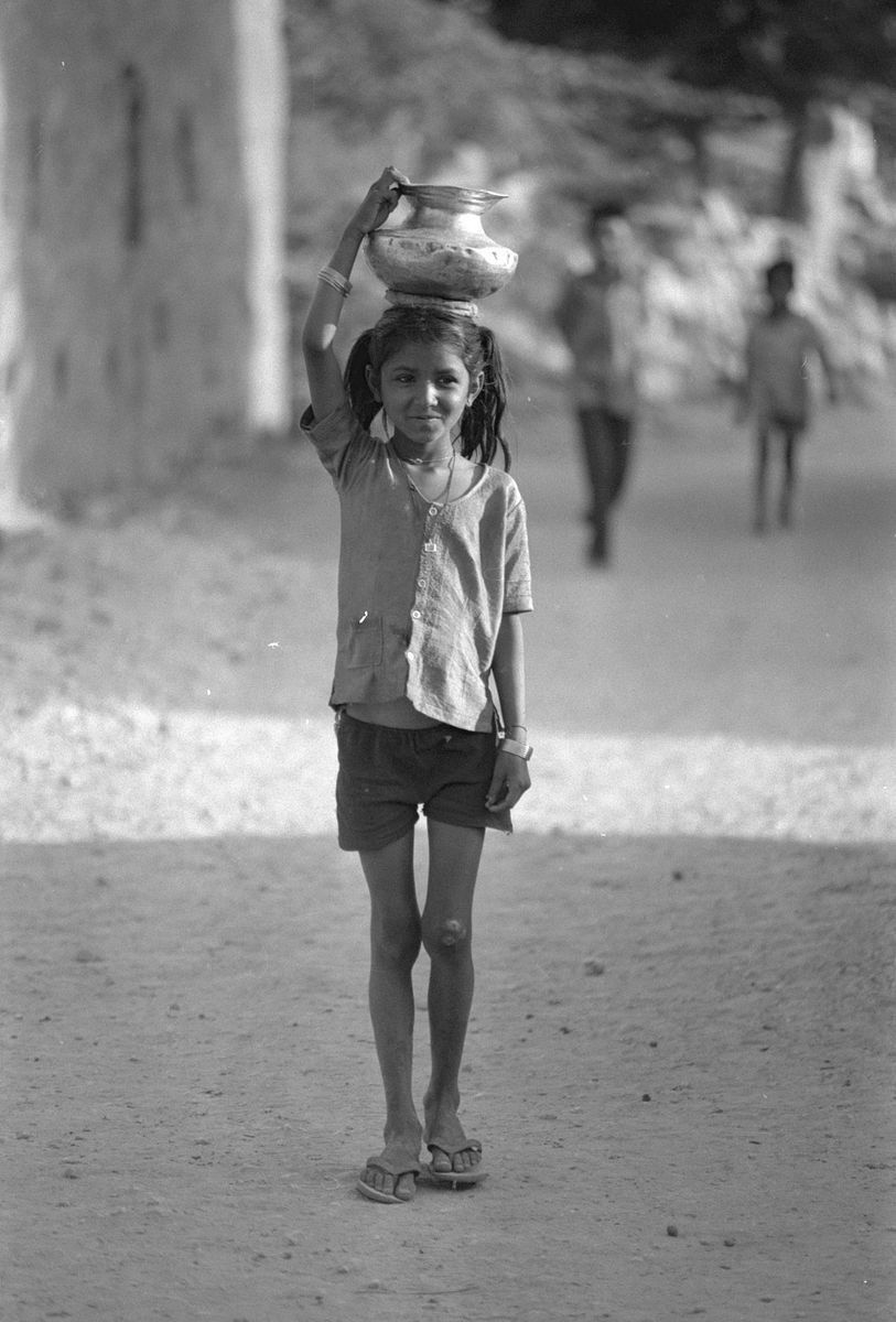 A smiling girl in flip-flops walking down a dusty path, balancing a metal pot on her head.
