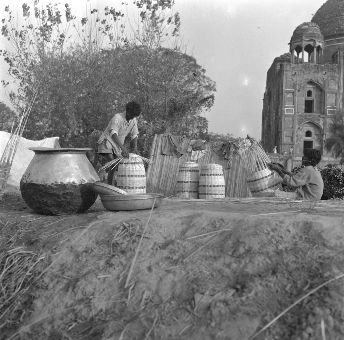 Men stacking woven baskets beside a large brass pot, with an old domed tomb behind them.