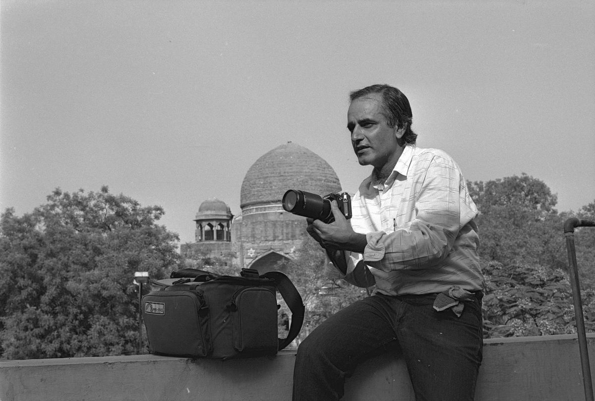Amar Talwar sitting on a wall with a camera and a long lens, his camera bag beside him and a domed tomb behind.